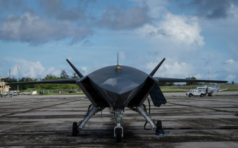 A dark, angular drone is parked on an airfield with a cloudy sky