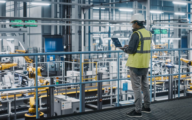 man with laptop looking out on robots on factory floor