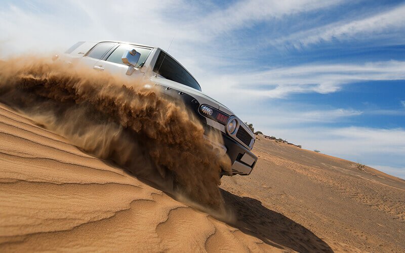 A white SUV drives through a sandy desert, kicking up a large cloud of dust.