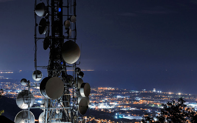 Image showing a nighttime shot of a large communications tower with multiple satellite dishes, overlooking a city skyline.