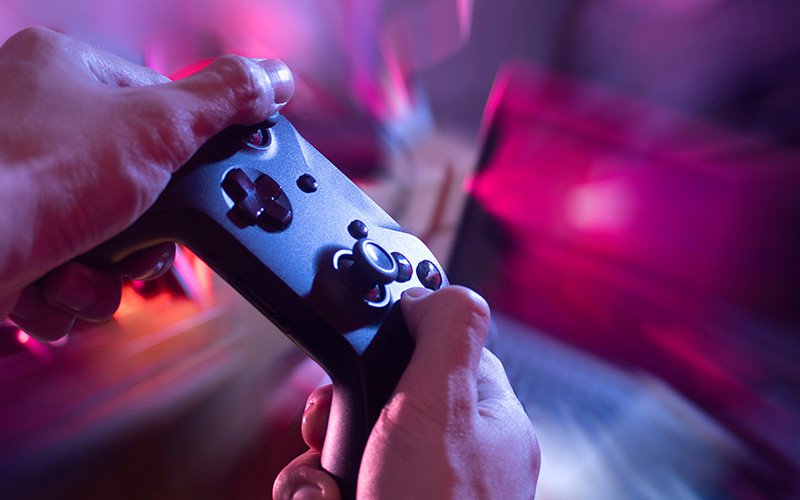 Hands hold a dark video game controller with a blur of pink and purple light in the background.