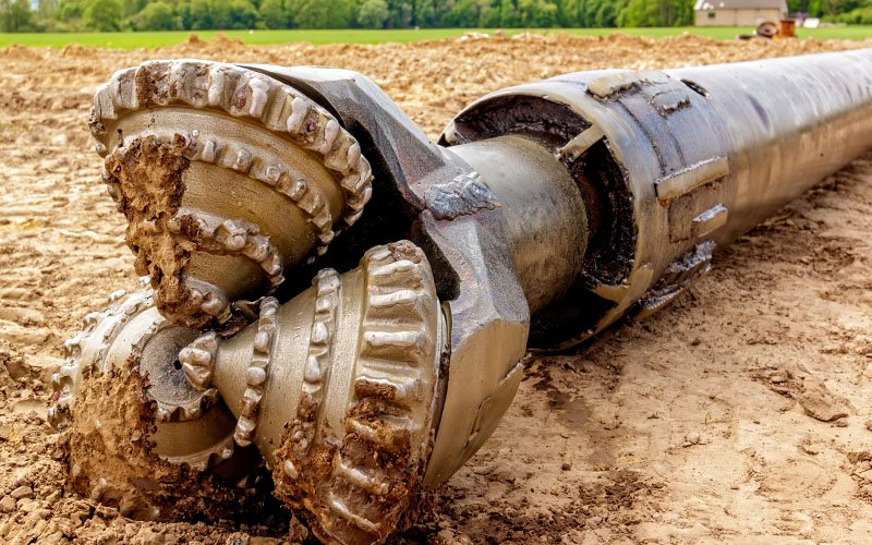 A close-up of a large drill bit covered in dirt lies on the ground.