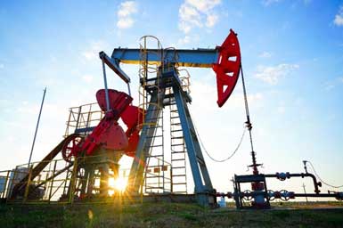 An oil pumpjack is silhouetted against a bright blue sky with a few white clouds.