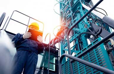 A worker in a blue uniform and yellow hard hat stands near a large drilling rig.