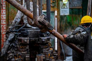 Workers in a drilling operation are seen handling a pipe with heavy machinery.